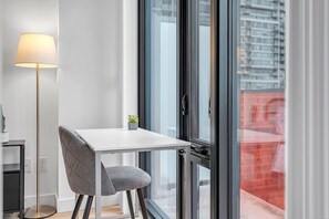 Habitació moderna amb una taula blanca, una cadira tapissada de color gris, una làmpada de peu i un finestral gran amb vista a edificis.