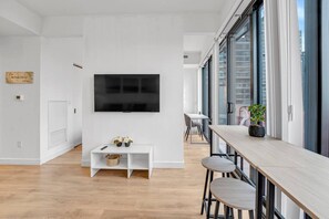 A modern living room with a flat-screen TV mounted on the wall, a white console table with decorative items, a dining area with a long table and chairs, and large windows offering a view of the city. - *2 Bed Condo* Near UofT/Rogers/Scotia/Hospital/Gym (Toronto)