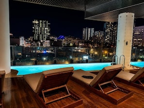 A rooftop pool area with lounge chairs and a view of the city skyline at night. - Central Fortitude Valley Stay | 1 Bed, 1 Bath (Fortitude Valley)