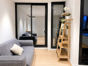 A modern living room with a grey sofa, a wooden ladder shelf with plants, and a television on a stand. - Central Fortitude Valley Stay | 1 Bed, 1 Bath (Fortitude Valley)