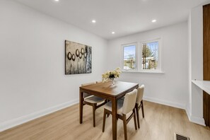 Een eetgedeelte met een houten tafel en stoelen, een vaas met bloemen, en een schilderij aan de muur.