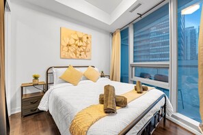 A hotel room with a large bed, a window with a view of buildings, a small bedside table with a plant, and a painting on the wall. - *10/10 Location* 2 BR Downtown Condo with GYM (Toronto)