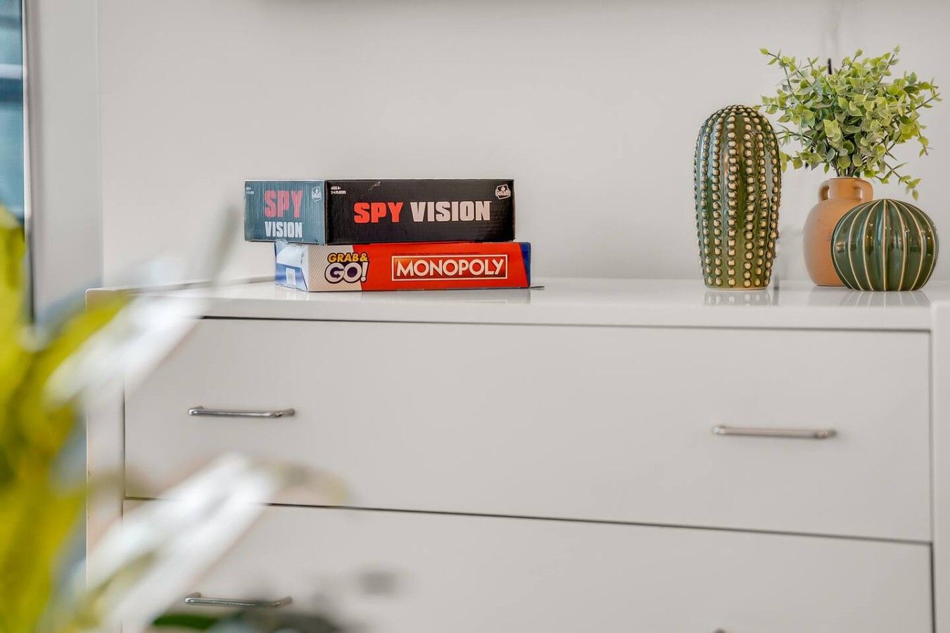 Un escritorio blanco con tres juegos de mesa: Spy Vision, Monopoly y un tercer juego. Hay dos jarrones decorativos con plantas sobre el escritorio.