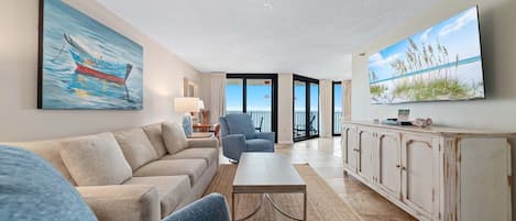 A living room with a sofa, armchair, coffee table, and a cabinet. There is a painting on the wall and a television screen displaying a beach scene.