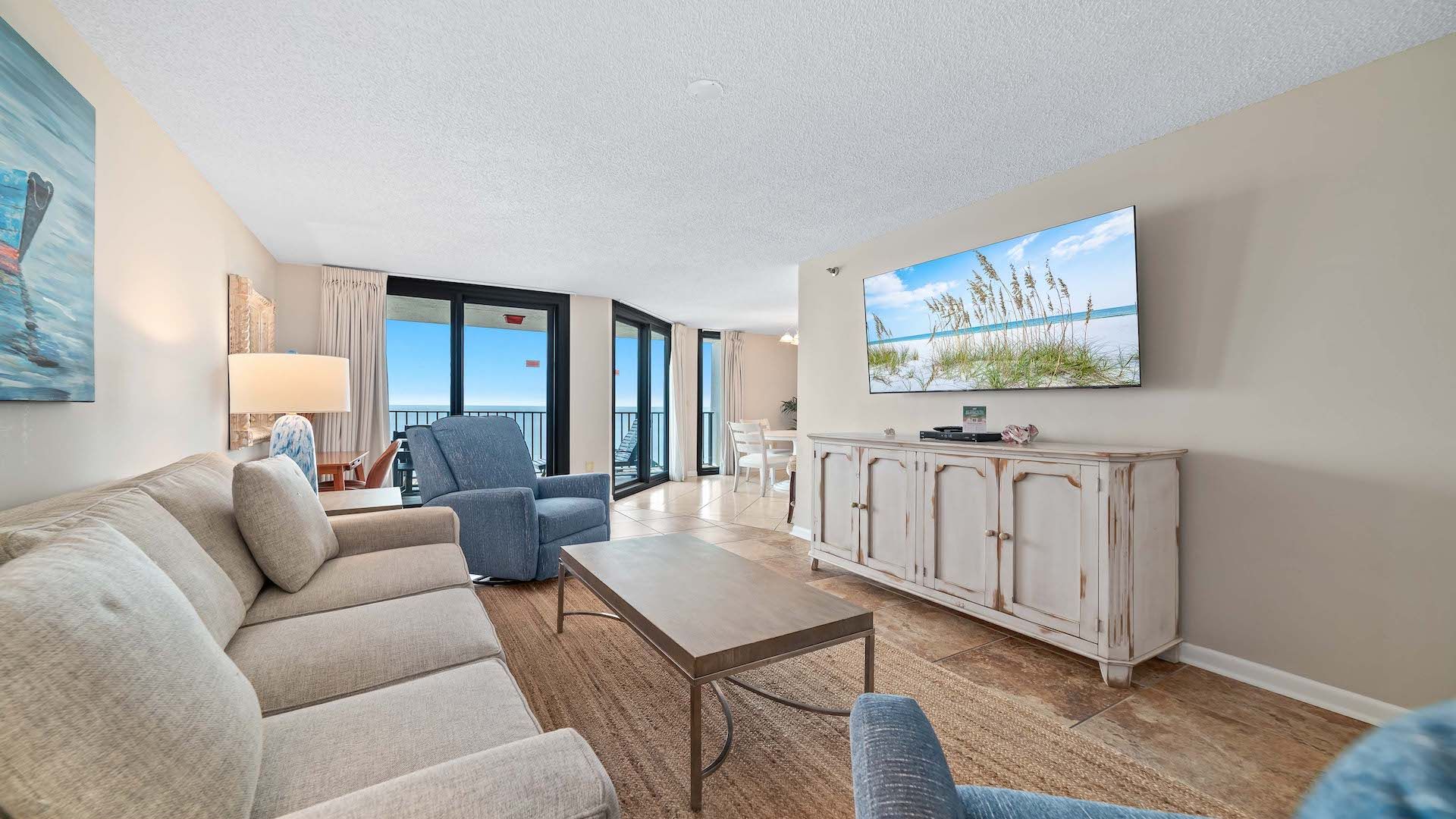 A living room with a sofa, armchairs, a coffee table, a TV, and a view of the beach through large windows.