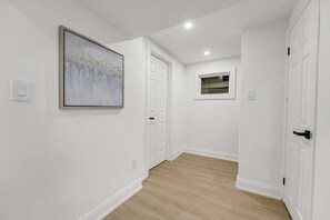 Un couloir aux murs blancs, doté d’un plancher en bois et orné d’un tableau abstrait encadré. On y trouve des portes blanches et une petite fenêtre donnant sur un paysage.