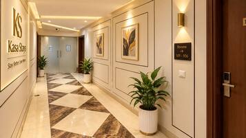 Een hotelgang met een geruit vloerkleed, potplanten, ingelijste kunstwerken en een bord met de kamer nummers 101-106.