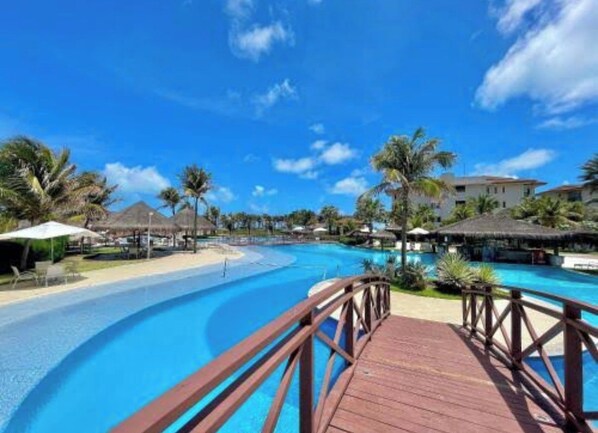 Área de piscina em resort com deck de madeira, palmeiras e construções com telhado de palha.