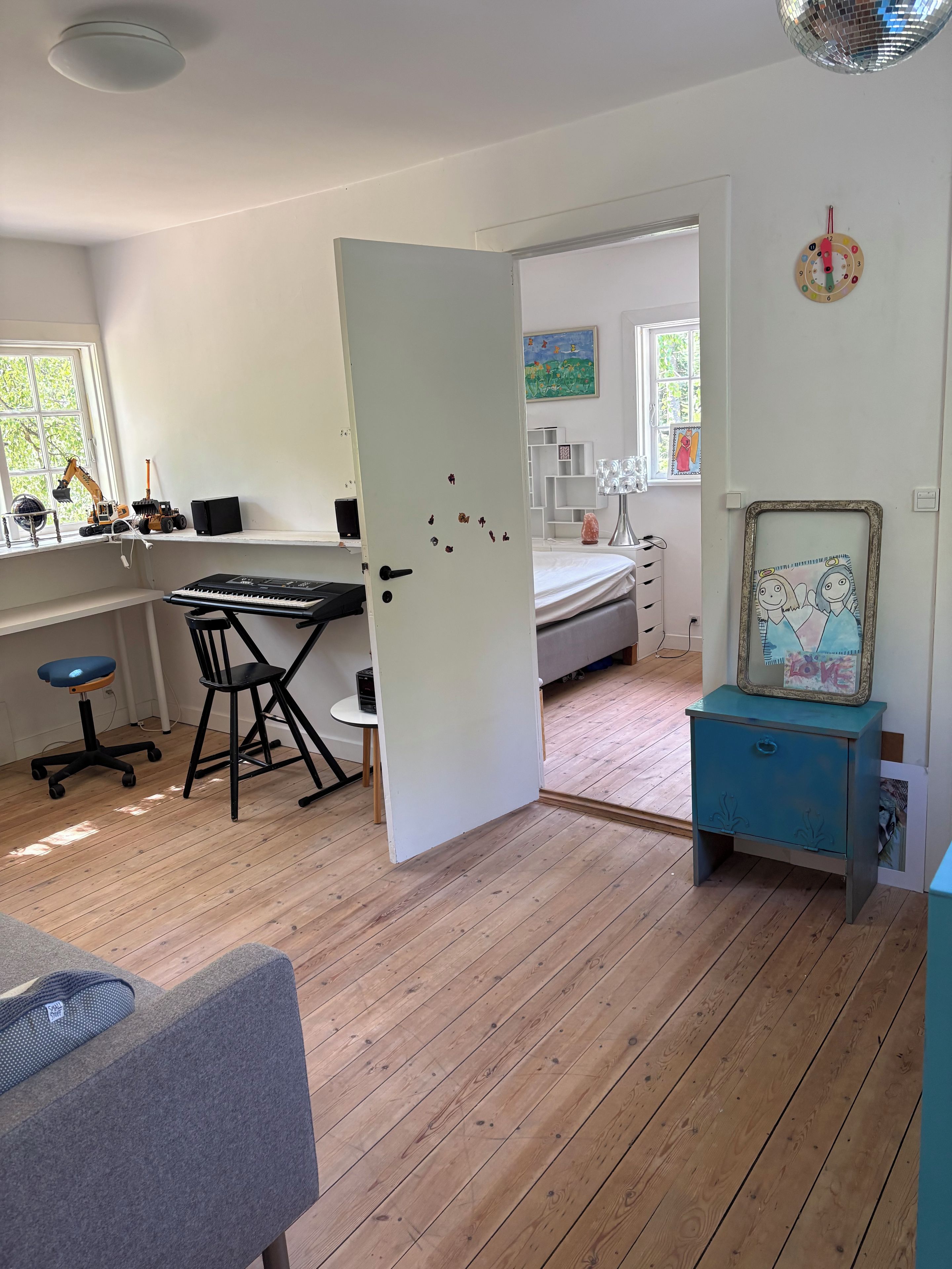 A room with a wooden floor, a desk with a chair, a bed, a small table with a picture frame, and a window with a view of trees.