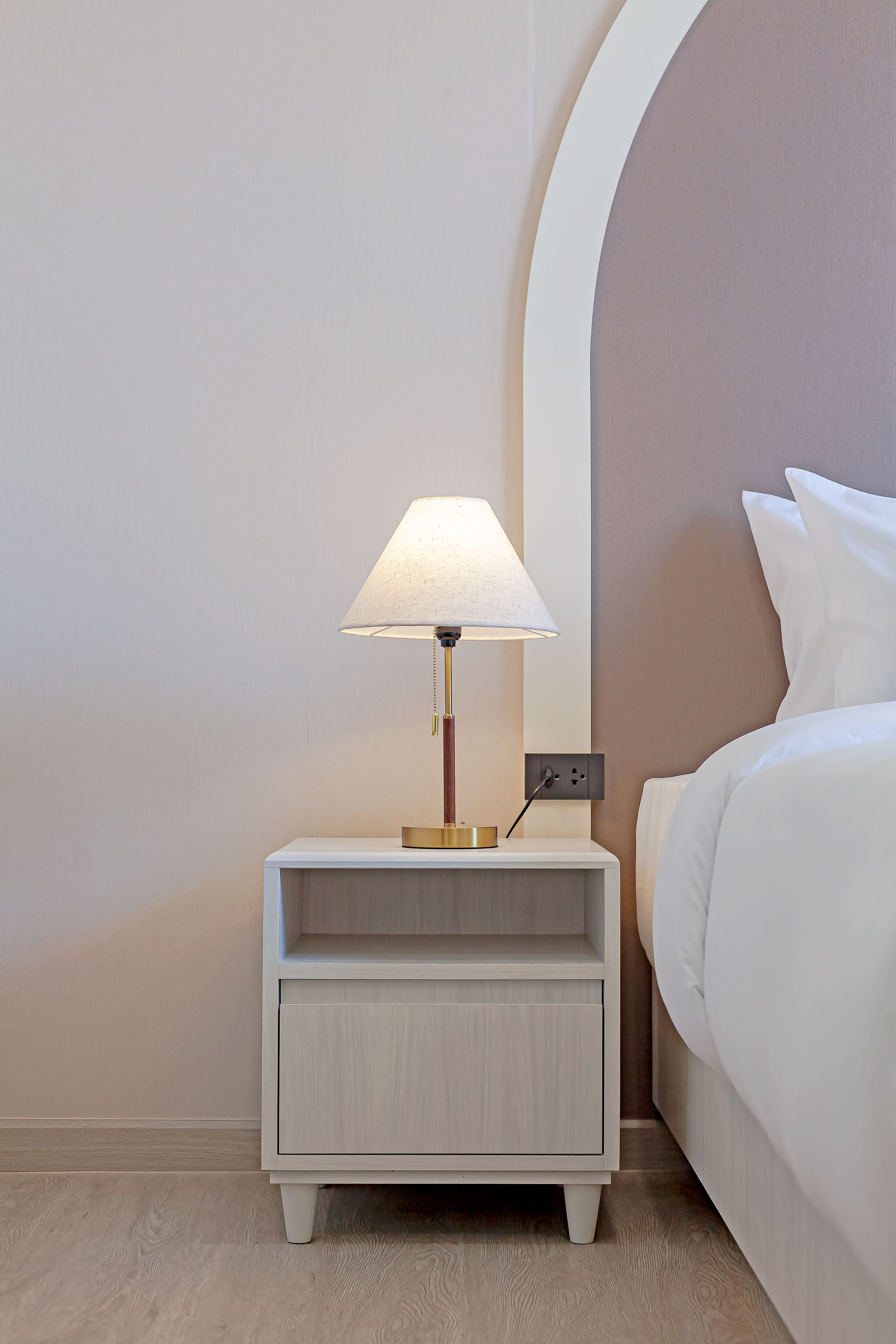 A neatly made bed with white linens, a bedside table with a lamp, and a power outlet on the wall.