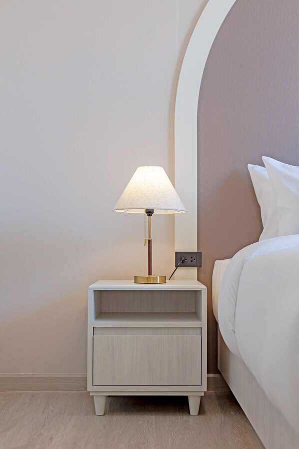 A neatly made bed with white linens, a bedside table with a lamp, and a power outlet on the wall.