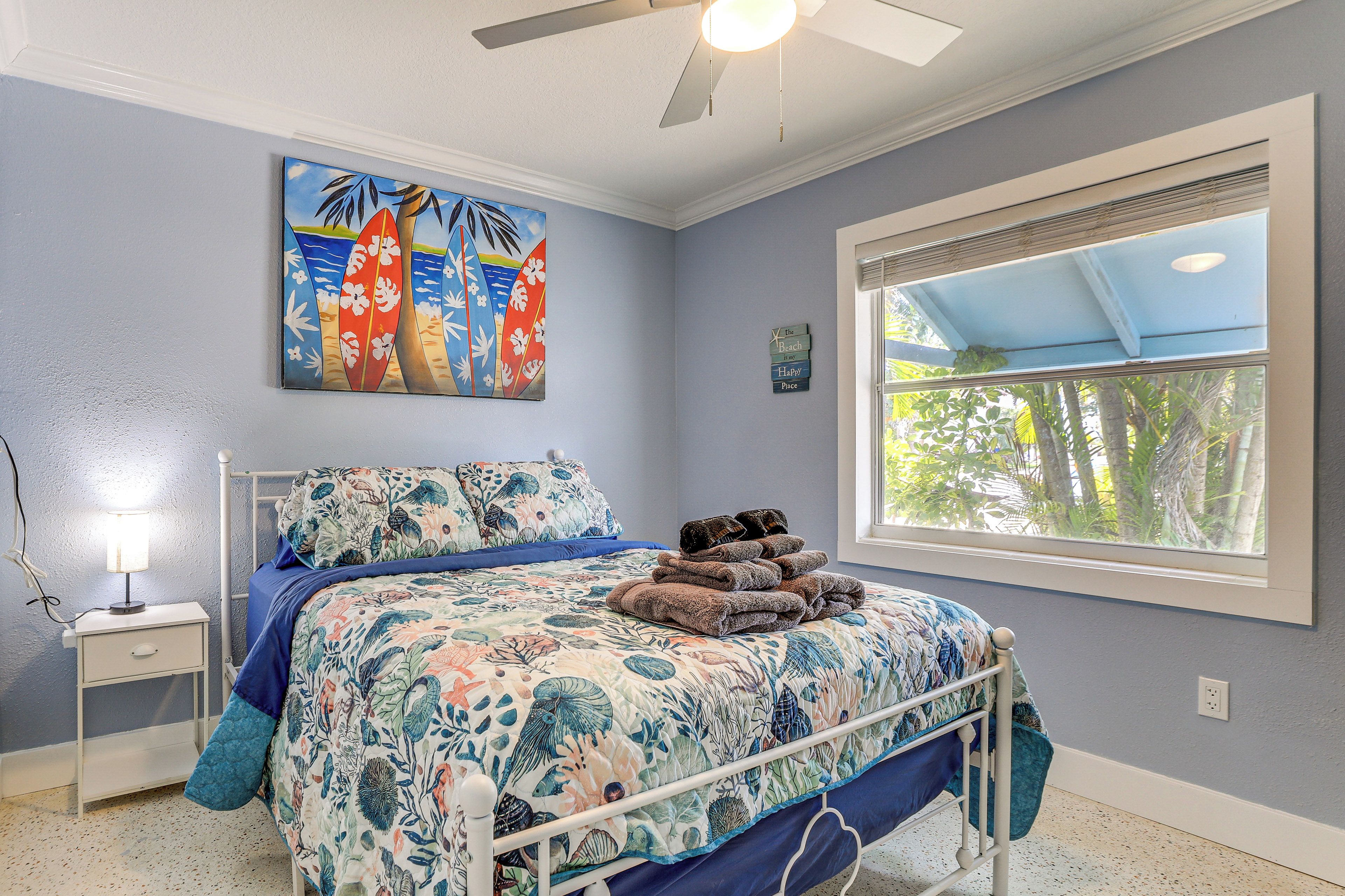A bedroom with a bed, a nightstand, a ceiling fan, a window with a view of trees, and a painting of surfboards.