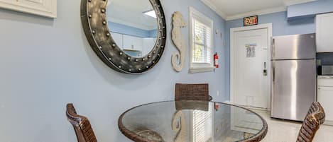 A dining area with a glass table and wicker chairs, a large mirror, and a kitchen with a stainless steel refrigerator.