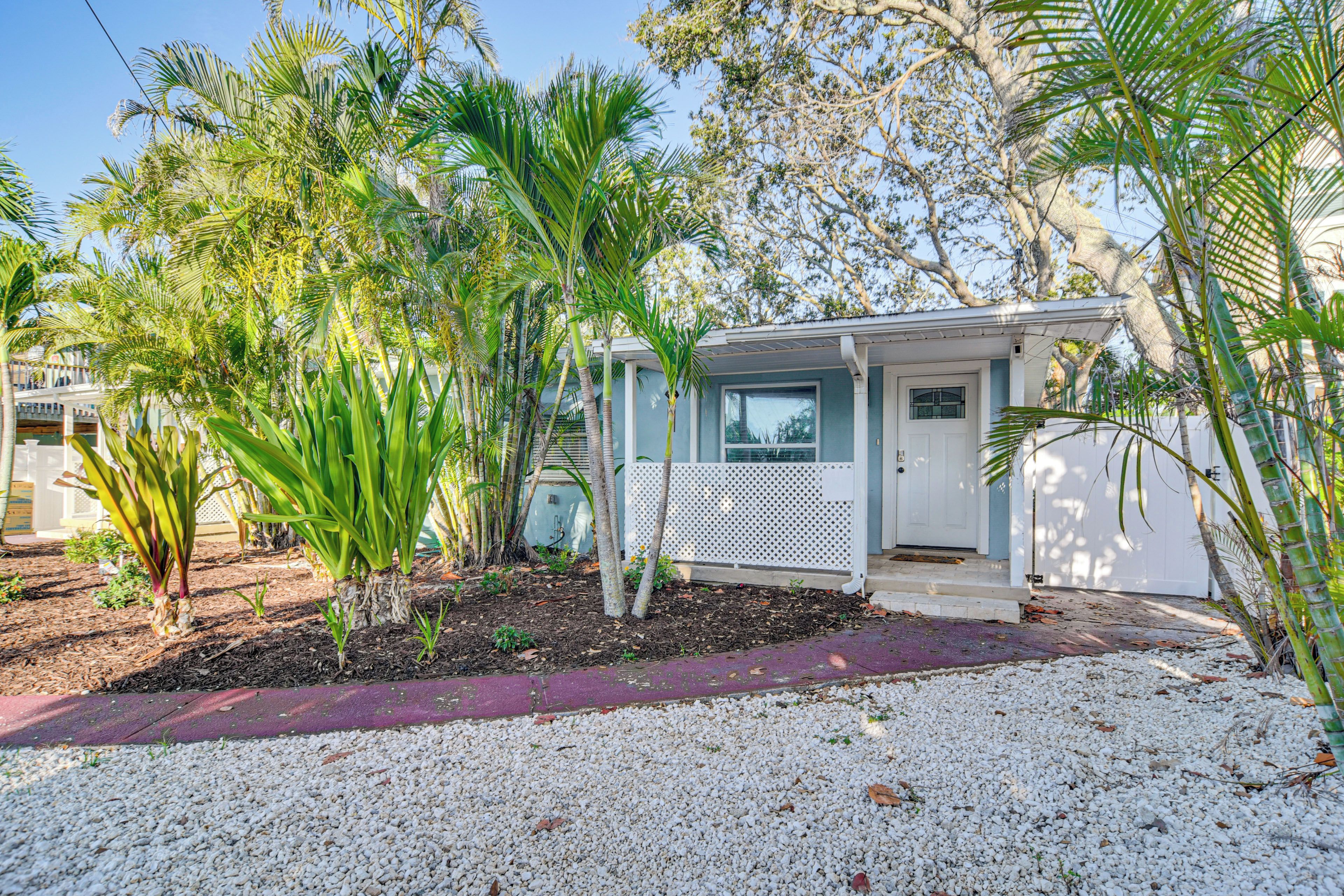 A small house with a porch, surrounded by palm trees and a gravel driveway.