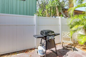 Ein tragbarer Grill, eine Propanflasche, ein weißer Klappstuhl und ein weißer Gartenzaun im Hinterhof.
