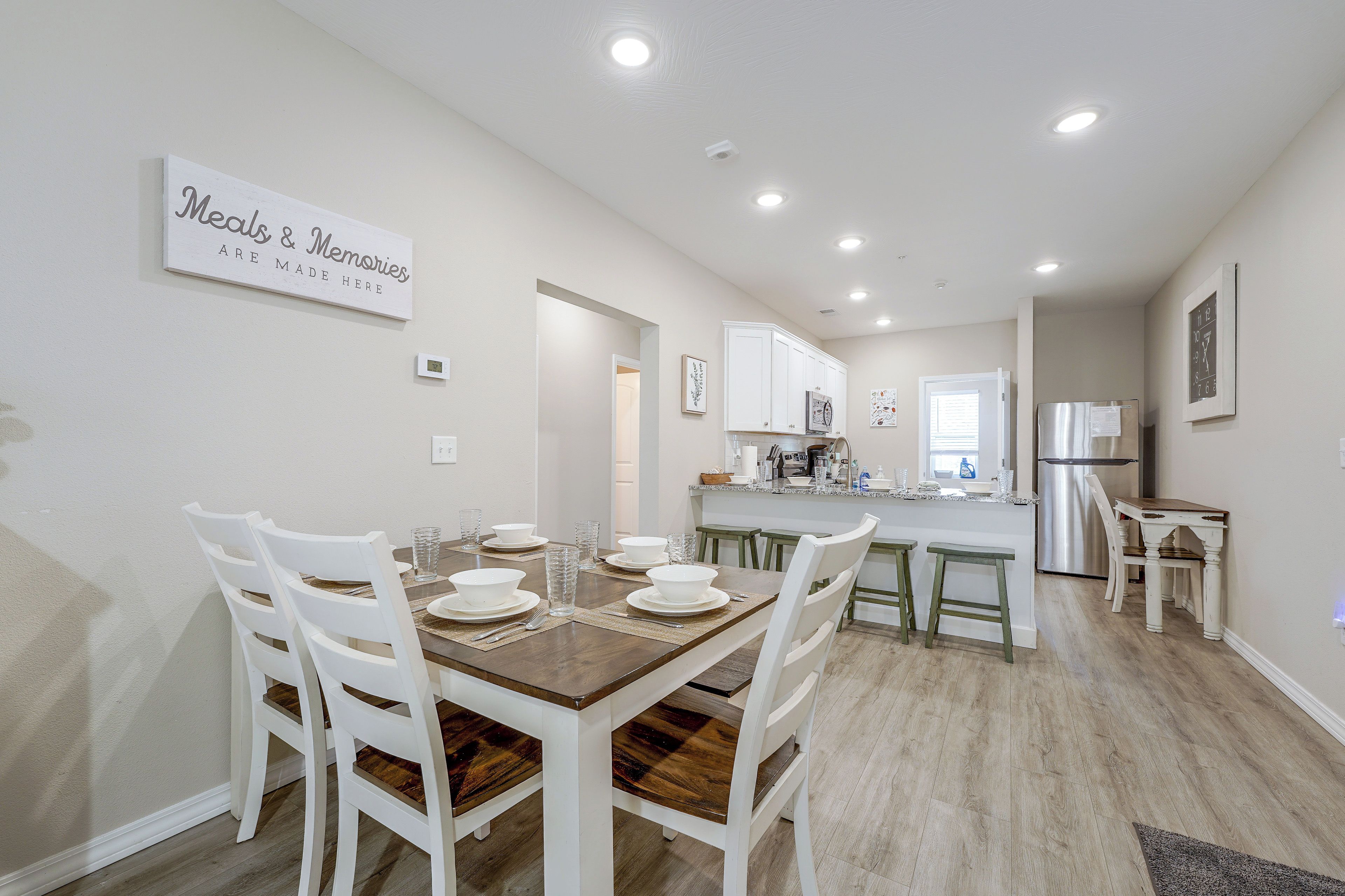A dining area with a wooden table and white chairs, a kitchen with white cabinets, and a sign on the wall.