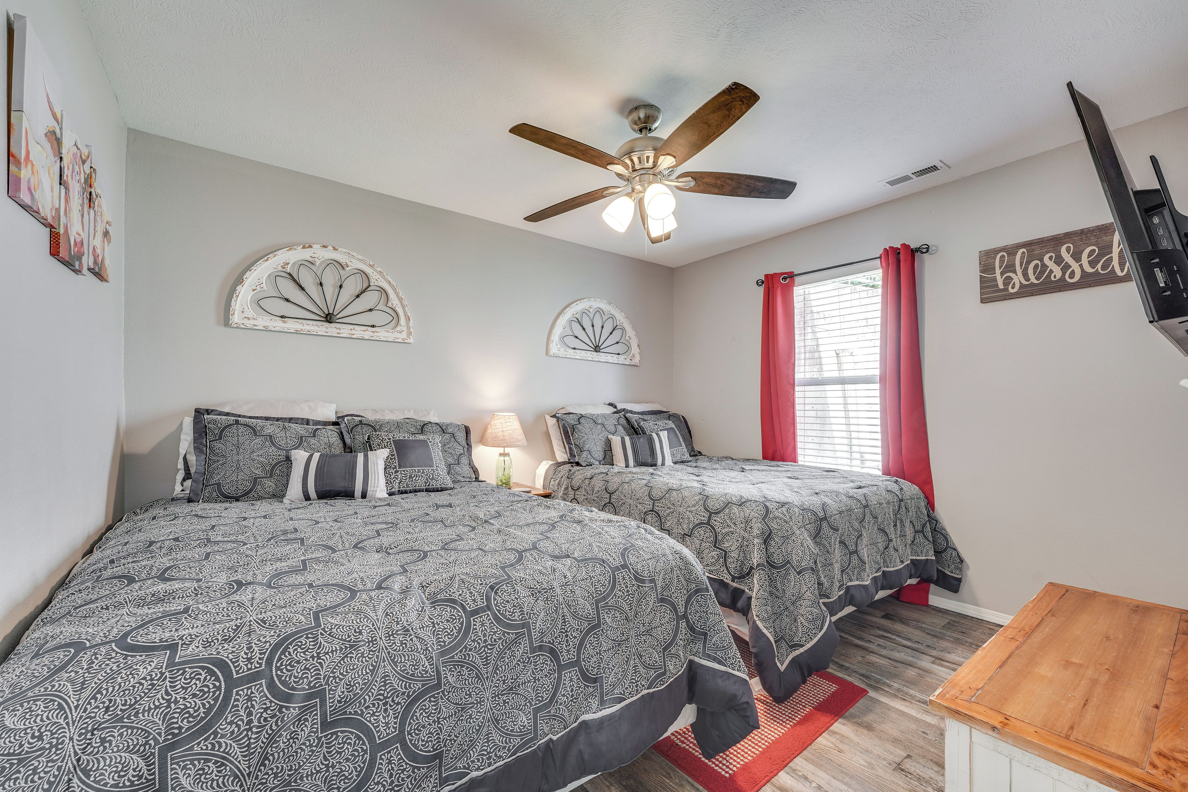 A bedroom with two beds, a ceiling fan, a window with red curtains, and wall art.