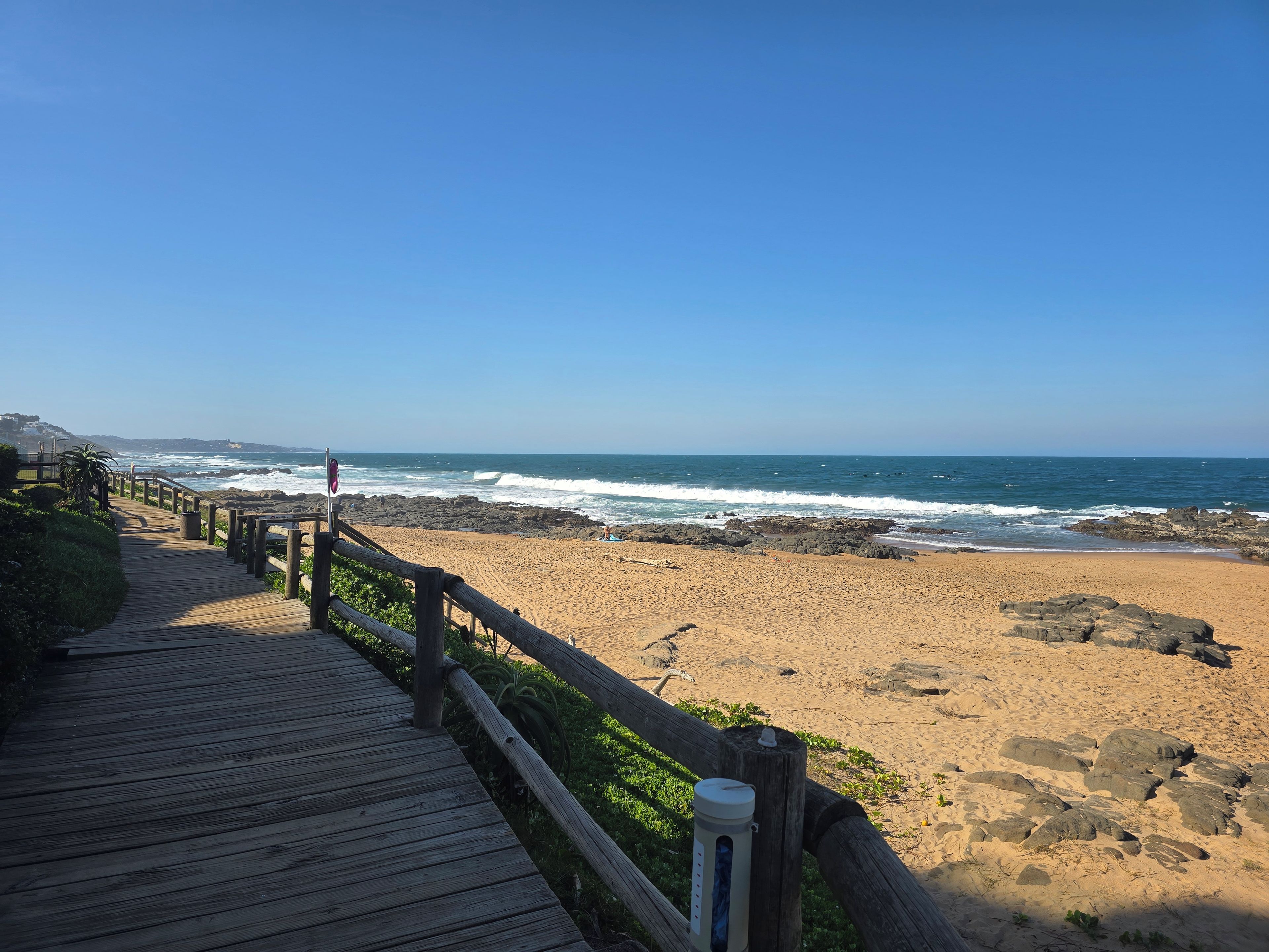 Un muelle de madera que conduce a una playa arenosa con afloramientos rocosos y el océano al fondo.