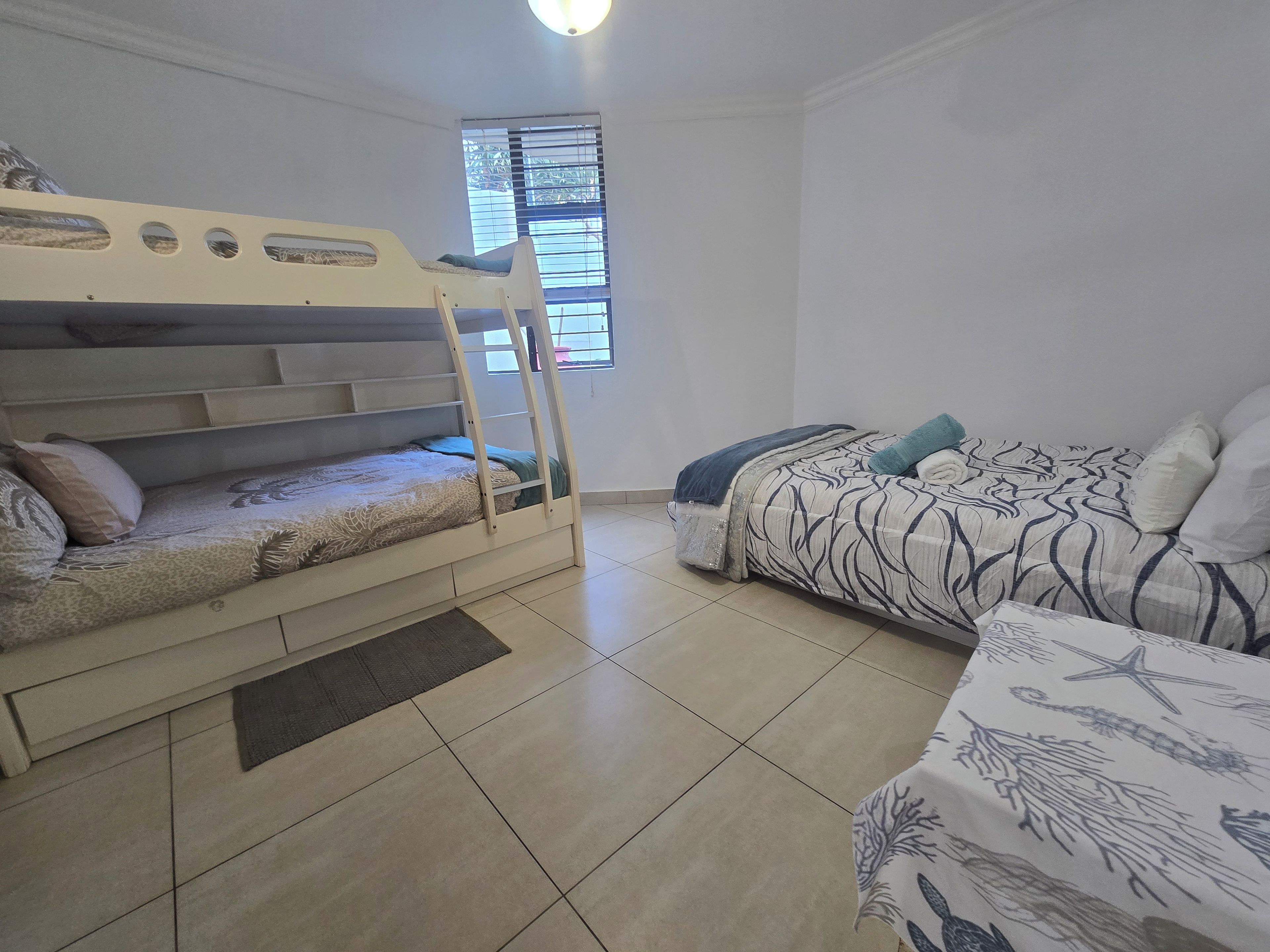 Een kamer met een stapelbed, een slaapbank, een raam met zonwering en een betegelde vloer.