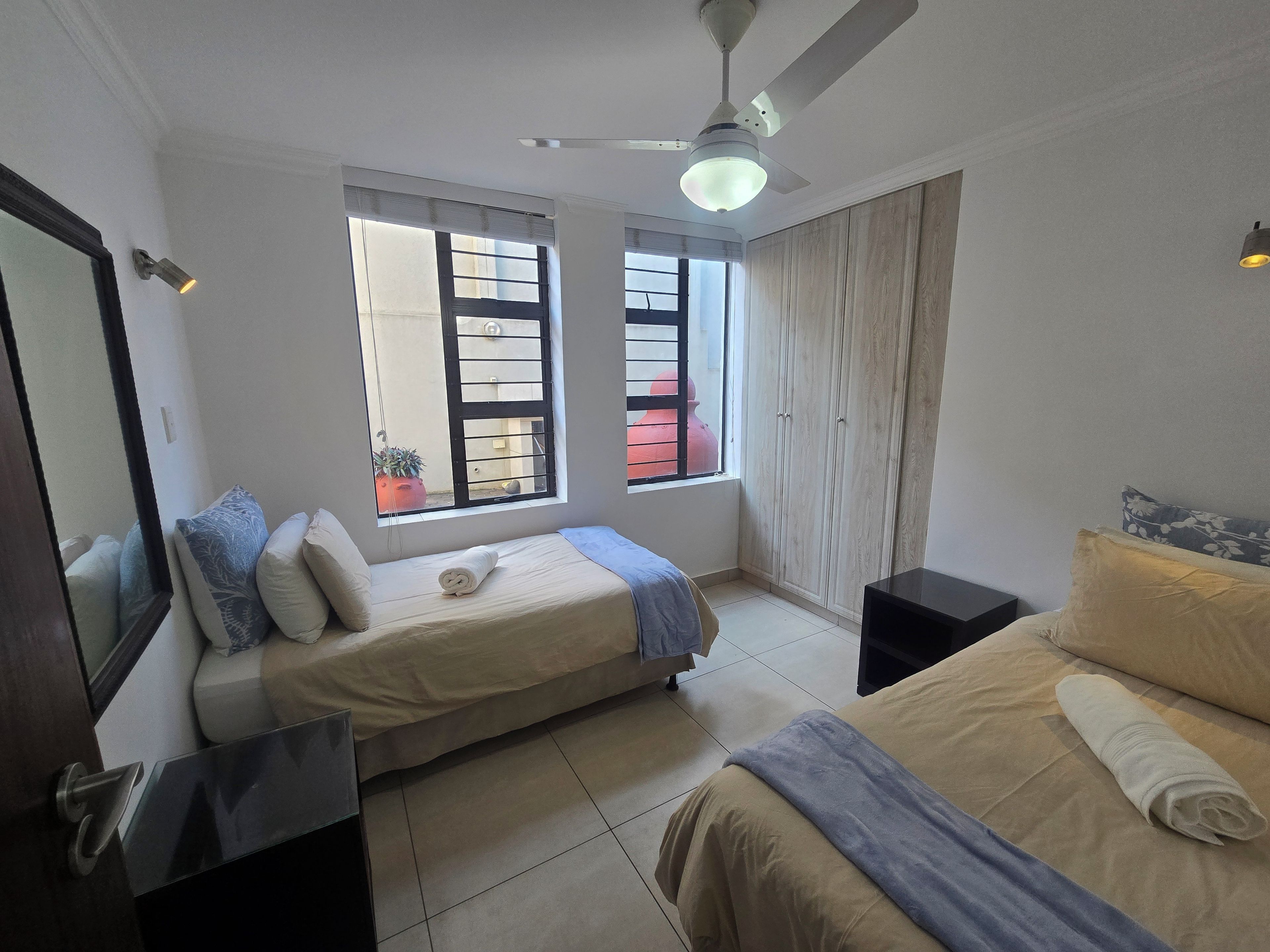 Un dormitorio con dos camas, ventilador de techo y ventanas amplias.