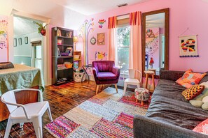 A cozy living room with a purple armchair, a sofa with colorful cushions, a bookshelf, and a large mirror.