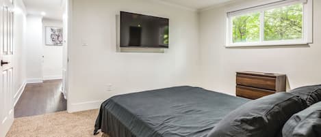 A bedroom with a bed, a dresser, a TV on the wall, and a window with a view of trees.