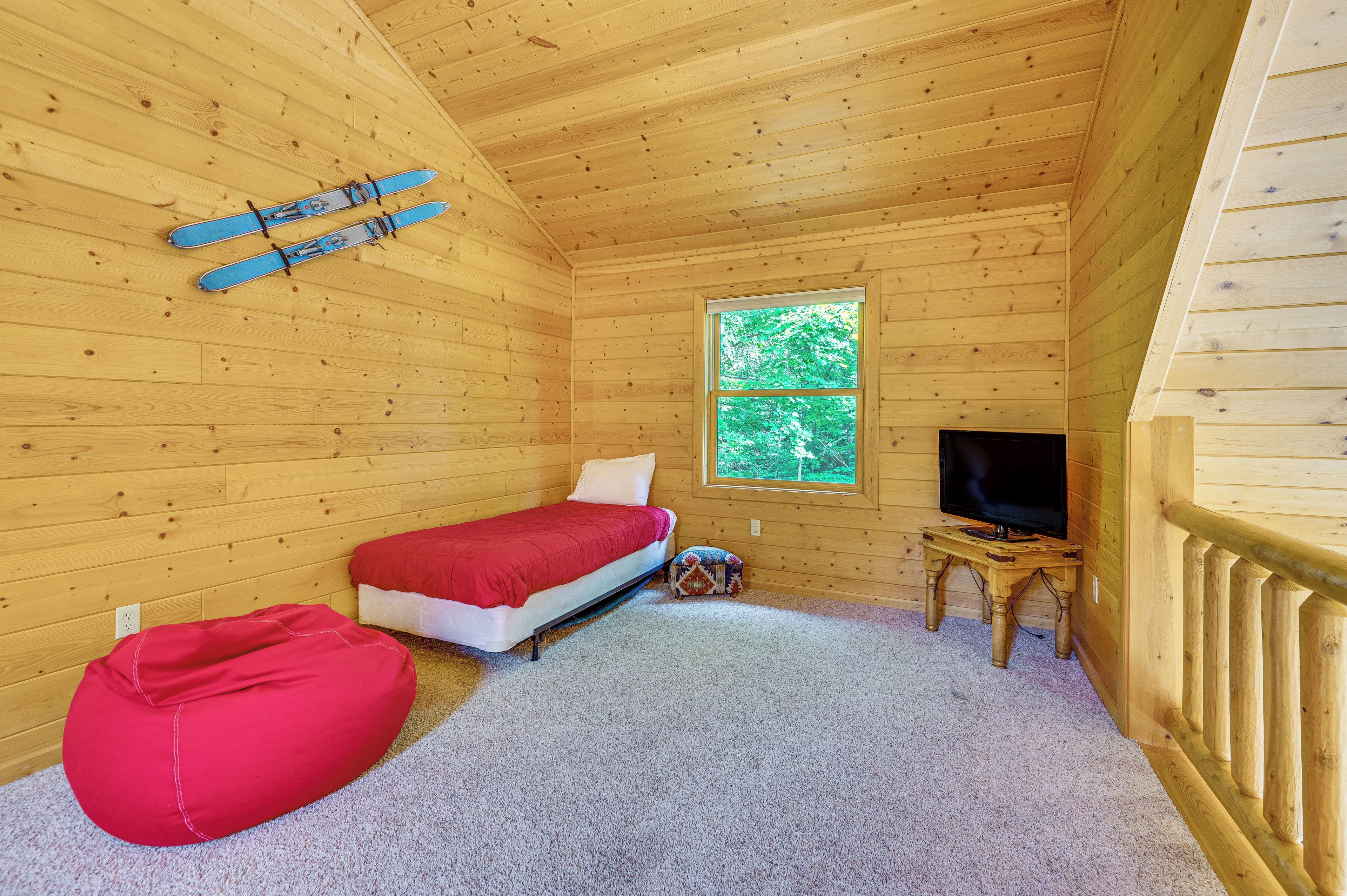 Une pièce de cabane en bois comprenant un lit, un téléviseur, une fenêtre et un pouf rouge.