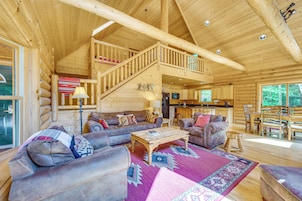 Interior espacioso de una cabaña de troncos con sala de estar, cocina y una escalera que lleva a un nivel superior.