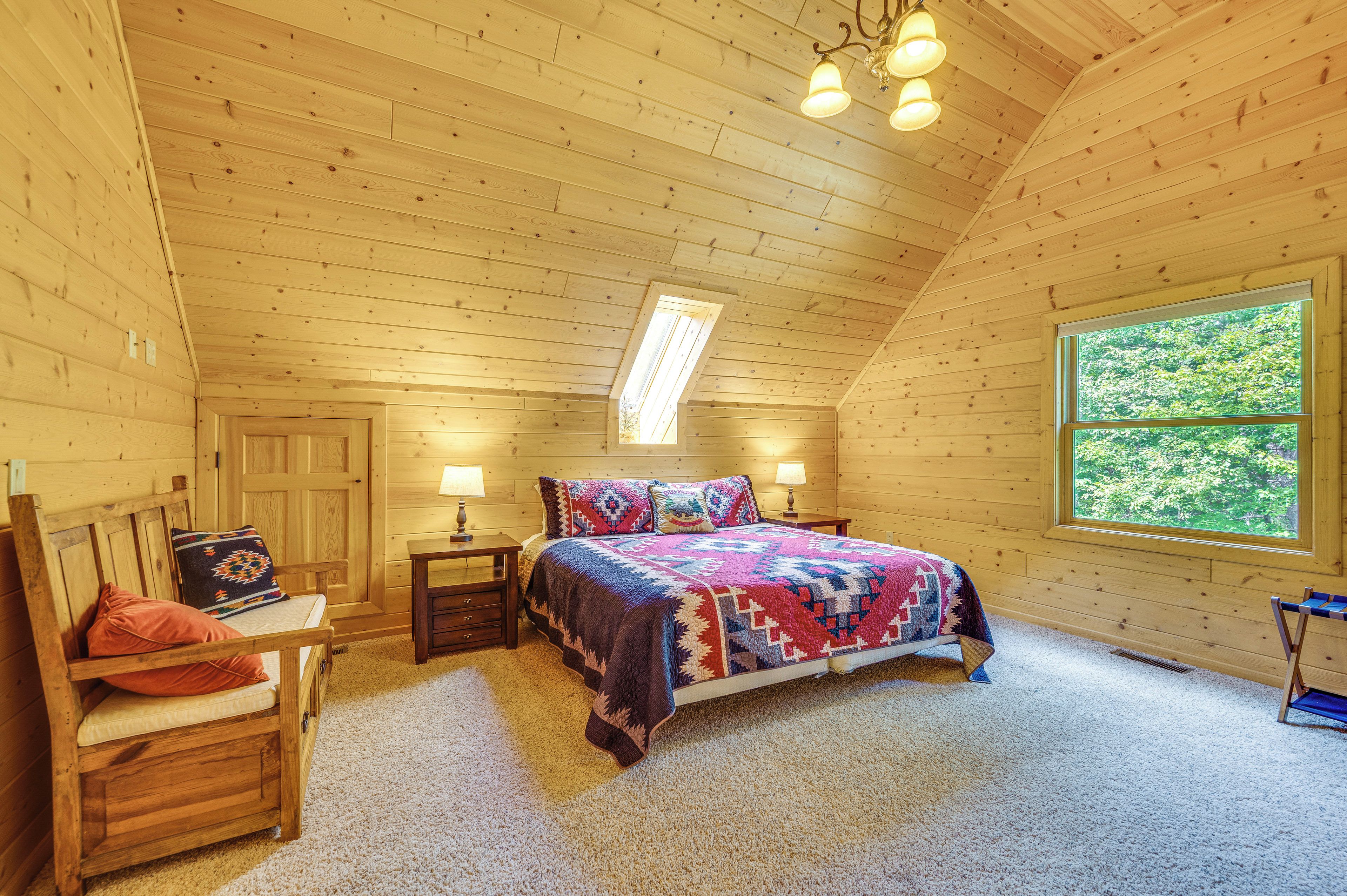 A cozy bedroom with a wooden ceiling, a large window, a bed with a colorful quilt, a nightstand, and a chair.