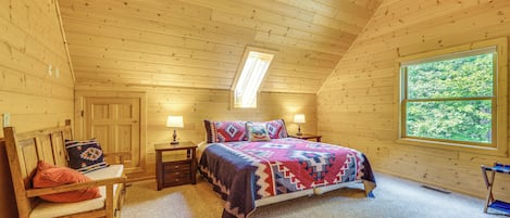 Un dormitorio acogedor con techo de madera, un ventanal grande, una cama con colcha colorida, una mesita de noche y una silla.