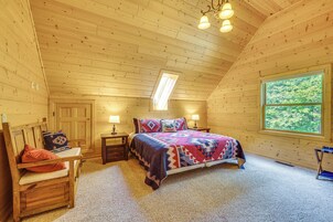 A cozy bedroom with a wooden ceiling, a large window, a bed with a colorful quilt, a nightstand, and a chair.