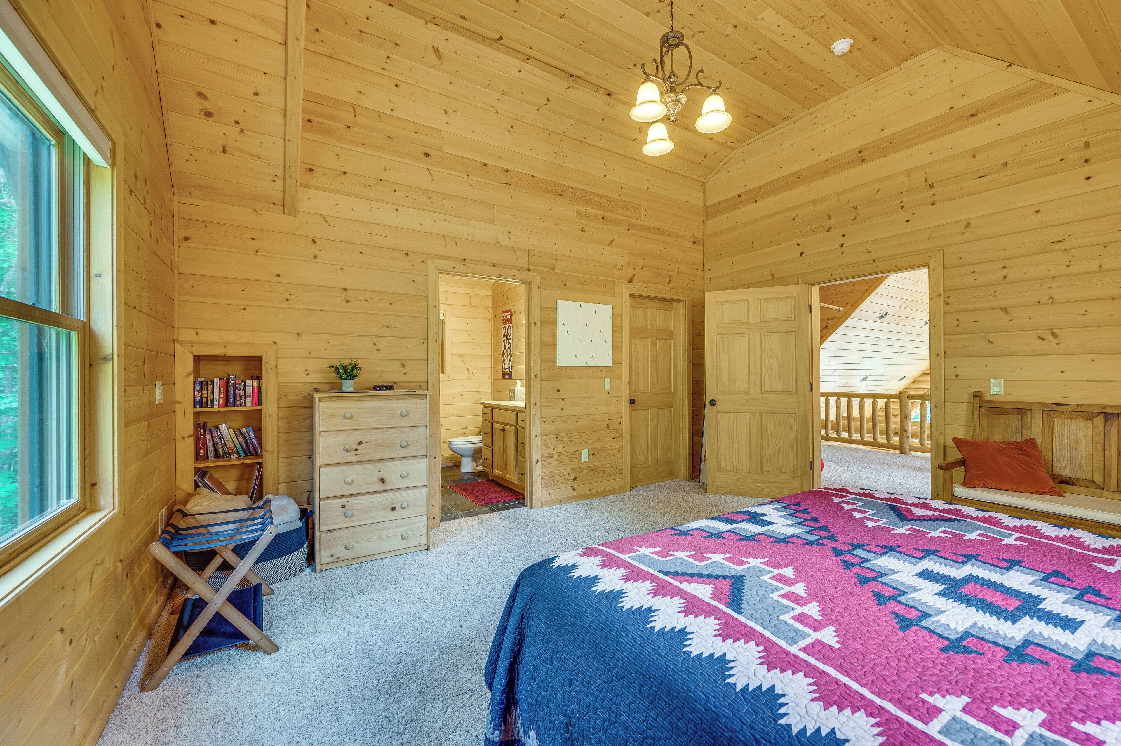 A wooden cabin bedroom with a bed, dresser, bookshelf, and a view of the outside.