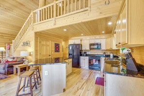 A wooden cabin interior with a kitchen, dining area, and a staircase leading to an upper level.