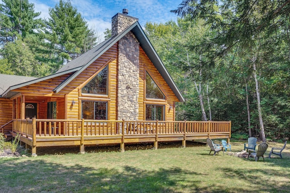 A log cabin with a stone chimney, wooden deck, and a view of the surrounding forest.