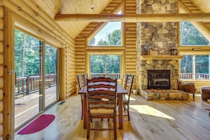 A log cabin interior with a dining area, stone fireplace, and large windows.