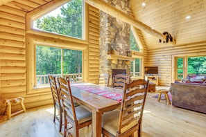 A log cabin interior with a dining area, stone fireplace, and large windows.