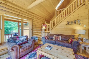 Interior de una cabaña de madera con una escalera de madera, dos sofás de cuero con cojines estampados, una mesa de centro de madera y vistas al exterior a través de amplias ventanas.
