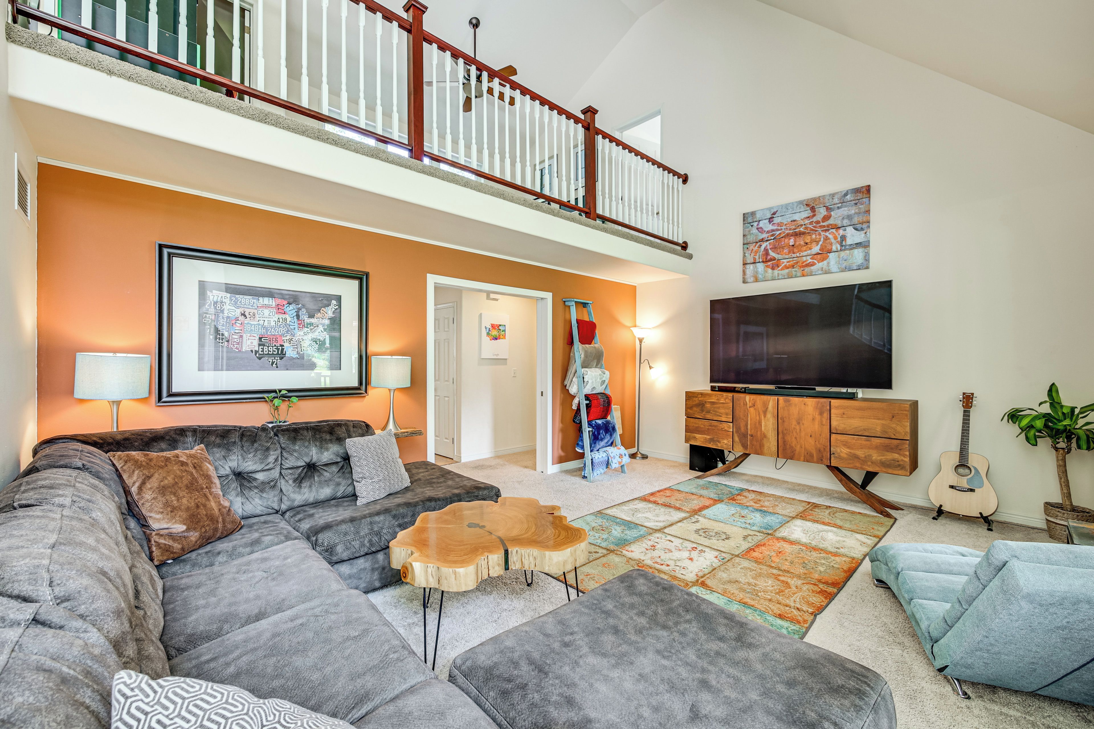 A spacious living room with a large sectional sofa, a wooden coffee table, and a colorful rug. A television is mounted on the wall, and there is a staircase leading to an upper level.