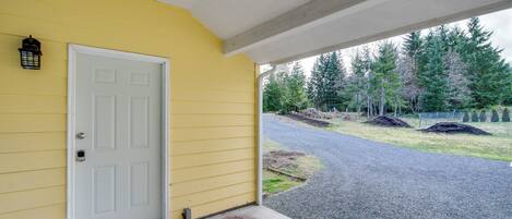 A yellow house with a white door, a porch, and a view of a gravel driveway and trees.