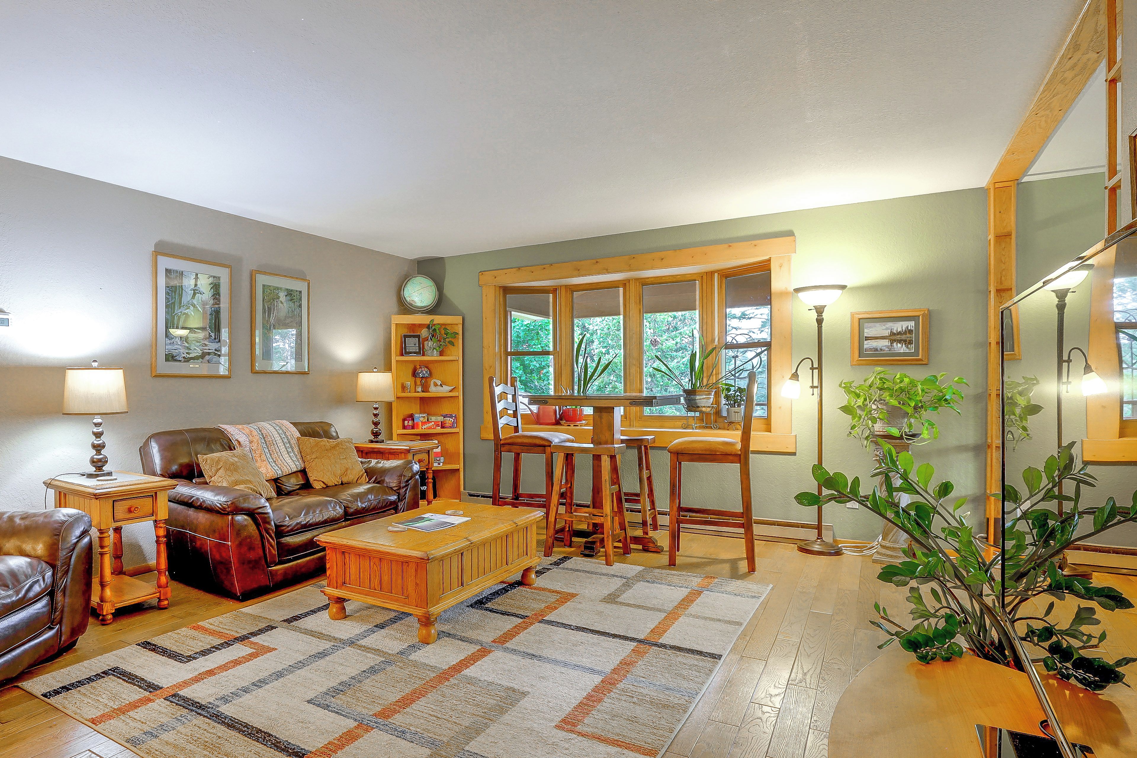 A living room with a brown leather sofa, wooden coffee table, and a dining area with wooden chairs and a table. There are framed pictures on the walls, a clock, and a potted plant.