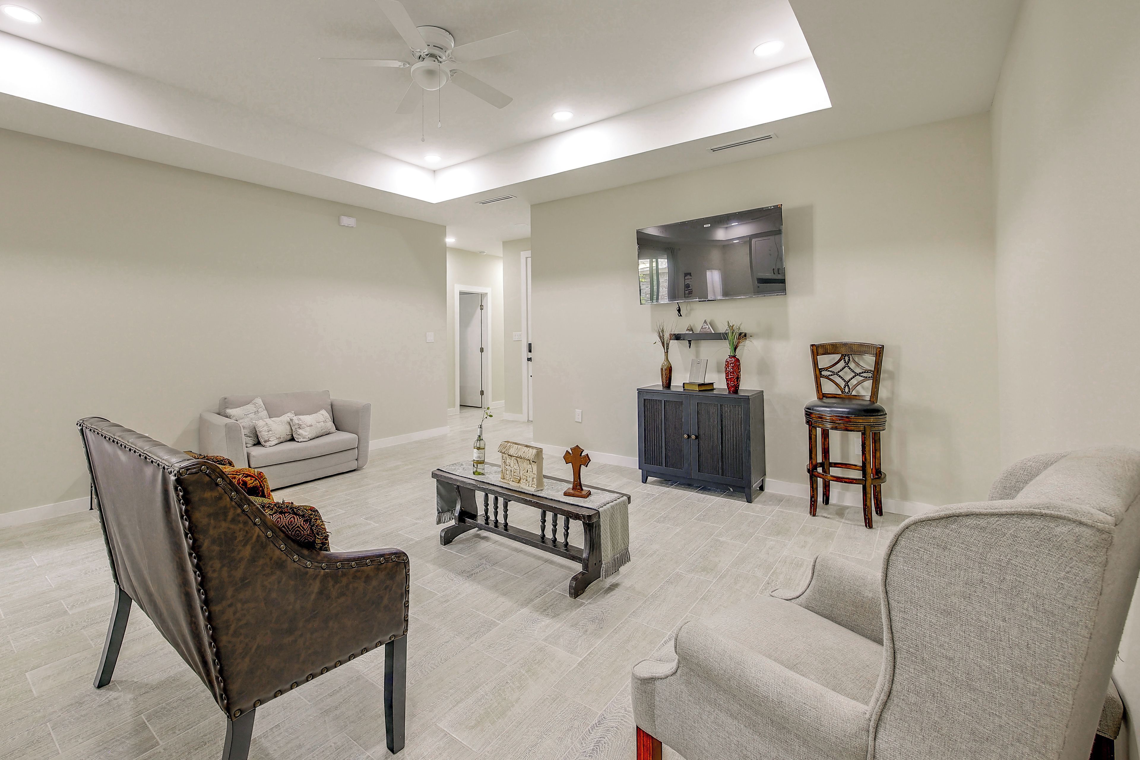 A spacious living room with a grey sofa, a dark leather chair, a coffee table, and a television mounted on the wall.