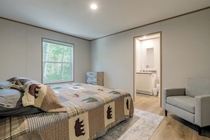 A bedroom with a large bed, a window with a view of trees, a bathroom with a mirror and sink, and a grey armchair.