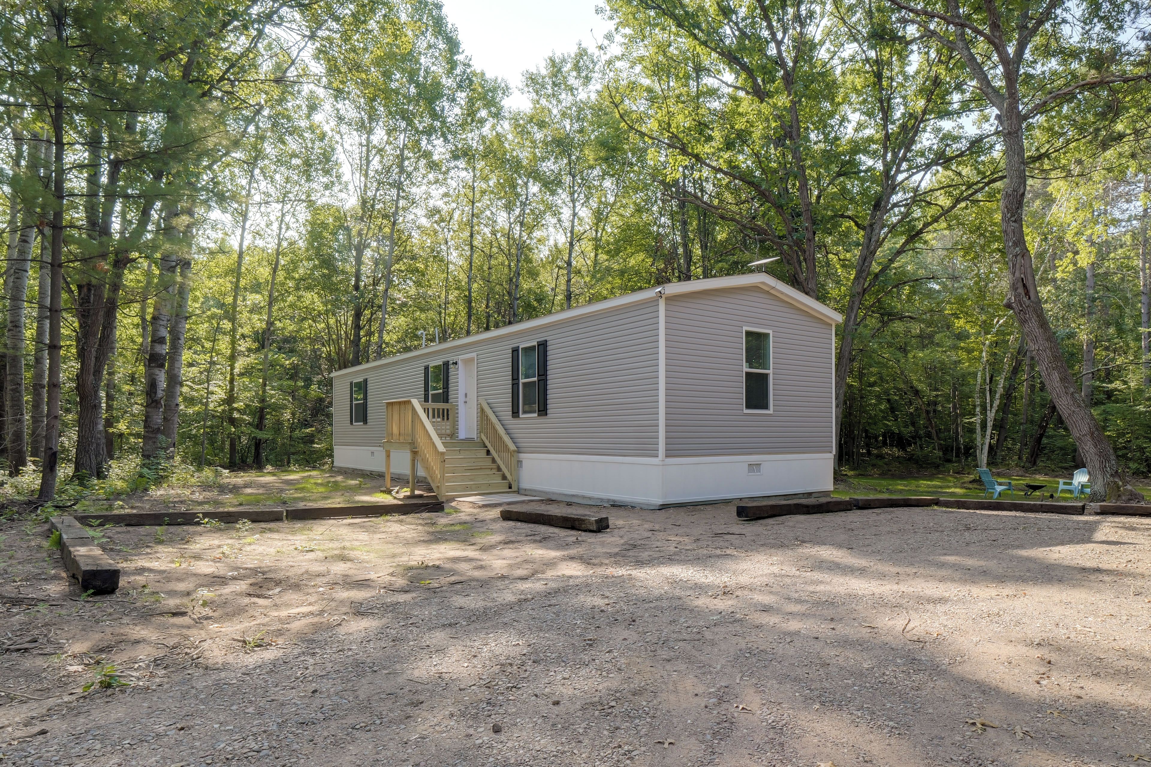 A single-story mobile home with a front porch and a small deck, surrounded by a wooded area.