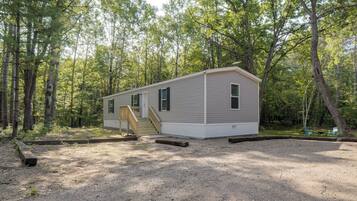 A single-story mobile home with a front porch and a small deck, surrounded by a wooded area.