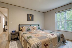 A bedroom with a bed, a nightstand, a digital clock, a window with a forest view, and a wall-mounted deer head artwork.