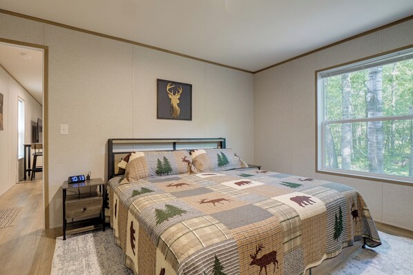 A bedroom with a bed, a nightstand, a digital clock, a window with a forest view, and a wall-mounted deer head artwork.