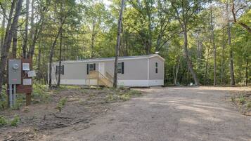 A single-story mobile home with a front porch and a small deck, surrounded by a forested area.