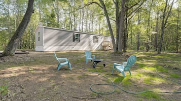 A mobile home in a wooded area with two outdoor chairs and a wheelbarrow.