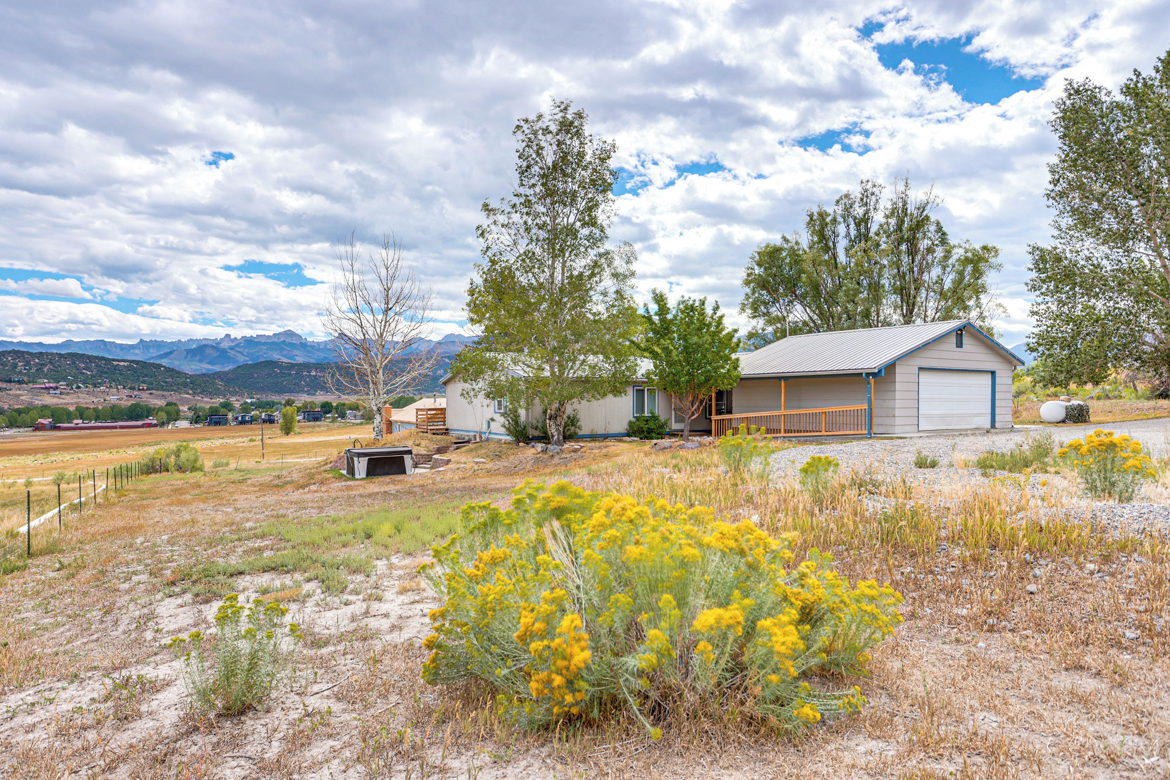 A rural house with a garage, surrounded by dry grassland and yellow flowers, with mountains in the background.