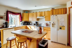 A kitchen with wooden cabinets, a refrigerator, a sink, and a window with a red curtain. - Walk to Yellowstone River! Emigrant Home w/ Patio (Emigrant)